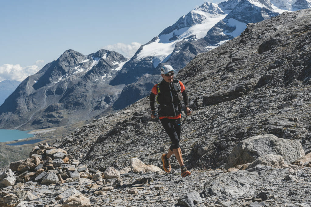 Trail runner on rocky trail with snowy peaks and a part of a lake at the background