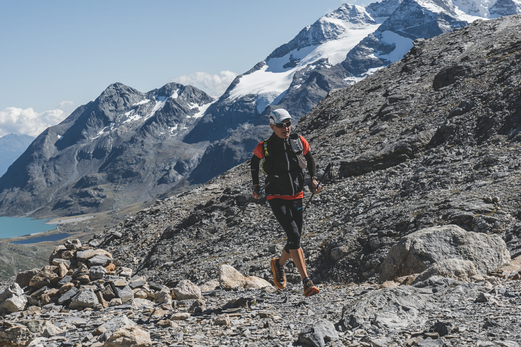 Trail runner on rocky trail with snowy peaks and a part of a lake at the background